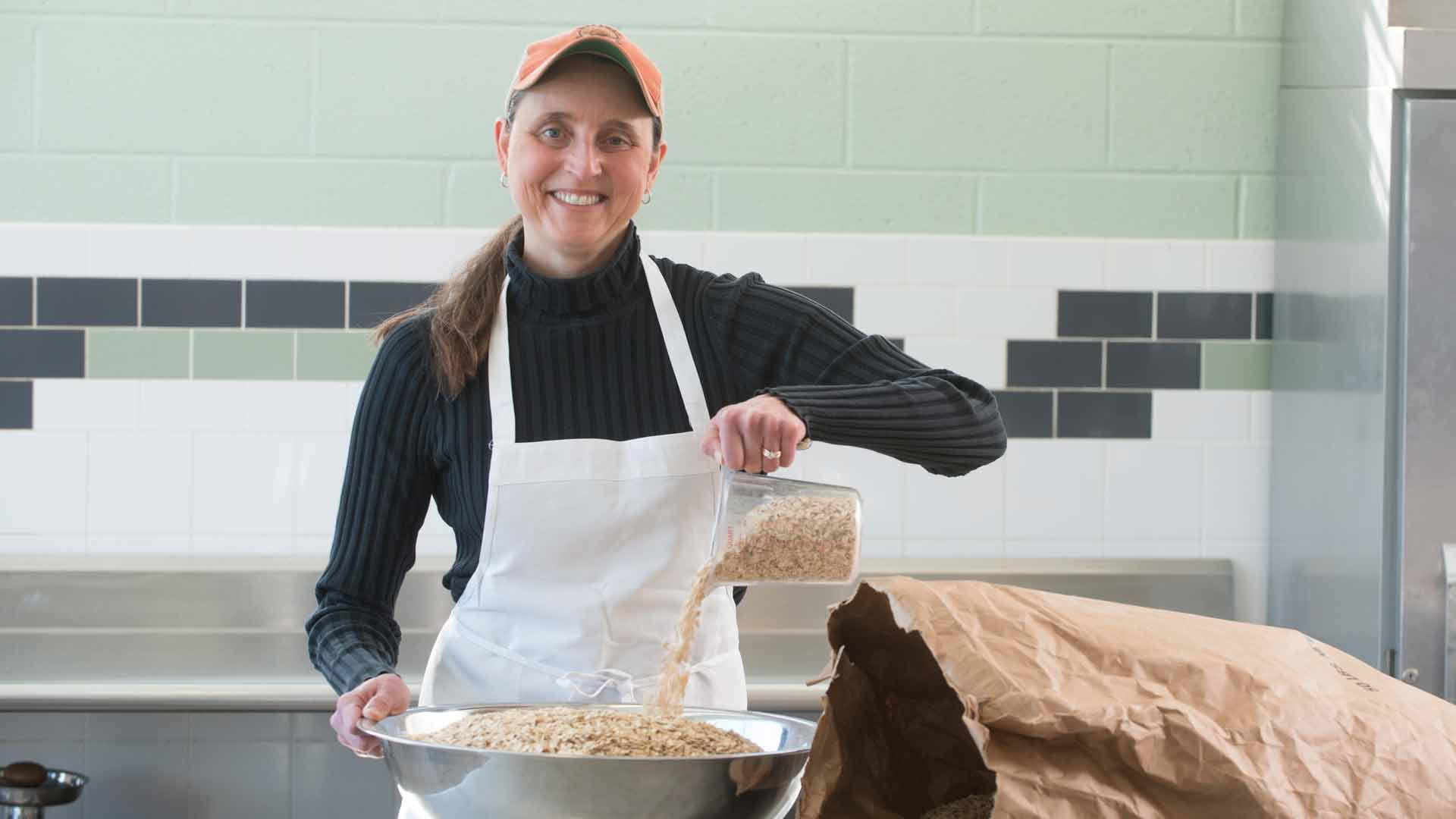 woman with apron on pouring granola into a large steel bowl