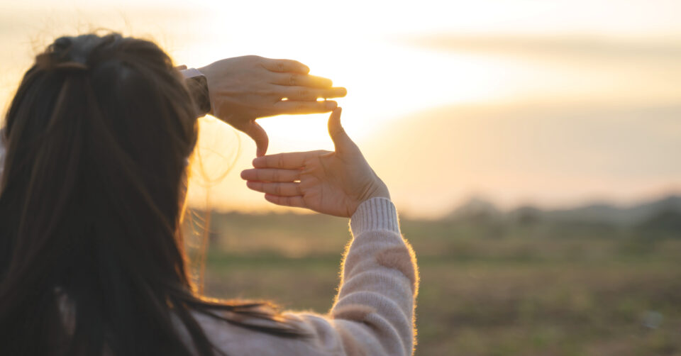 Back view of a person holding up their hands to frame the setting sun over a blurred field