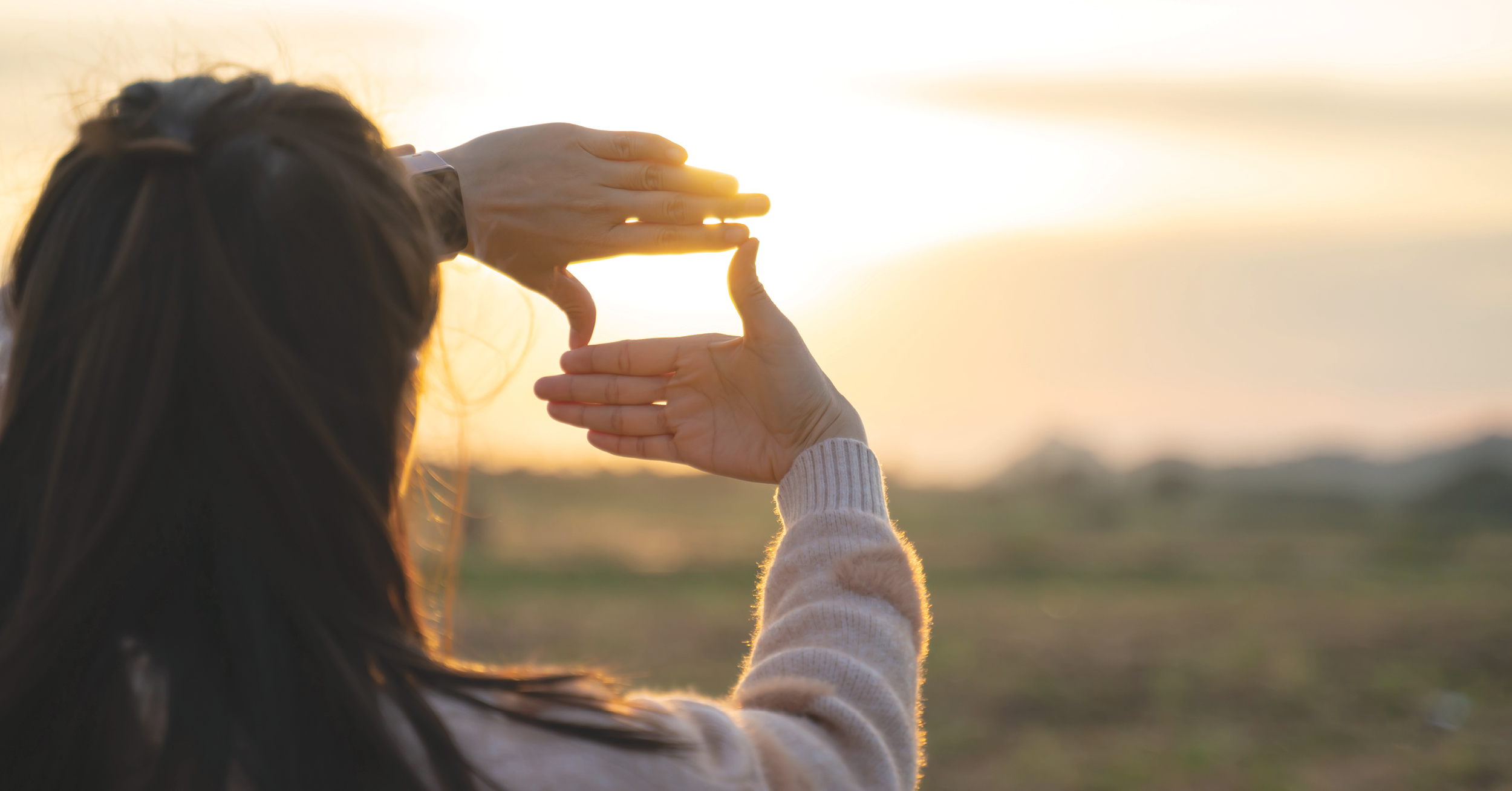 Back view of a person holding up their hands to frame the setting sun over a blurred field